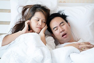 Woman in bed angrily covering her ears next to a snoring man
