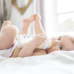 A baby on a white blanket drinking from a bottle 