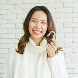 woman in white sweater holding Invisalign aligner in front of white brick wall 